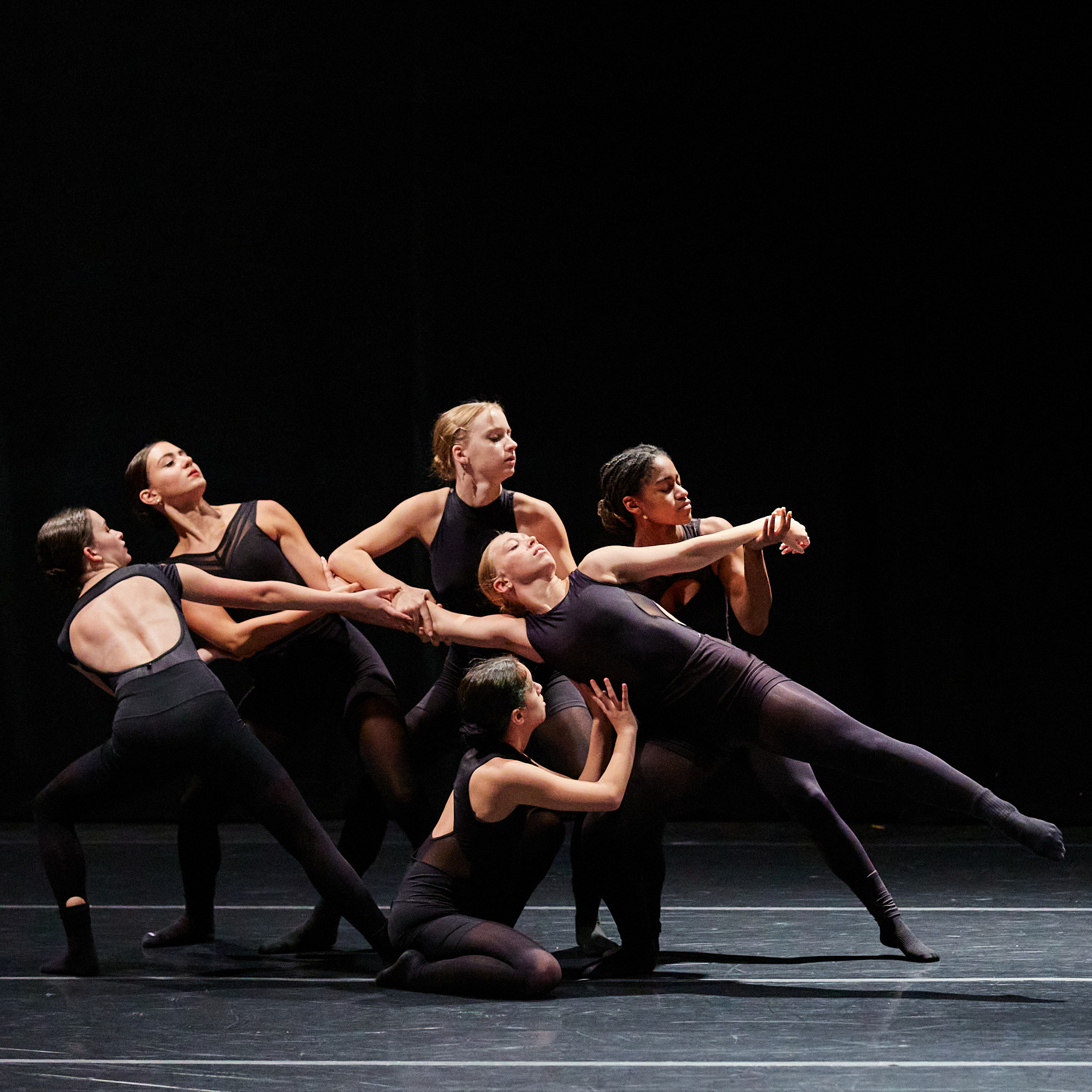 A group of six teen female dancers performing in black costumes. Five dancers are supporting a sixth dancer as she falls backwards.