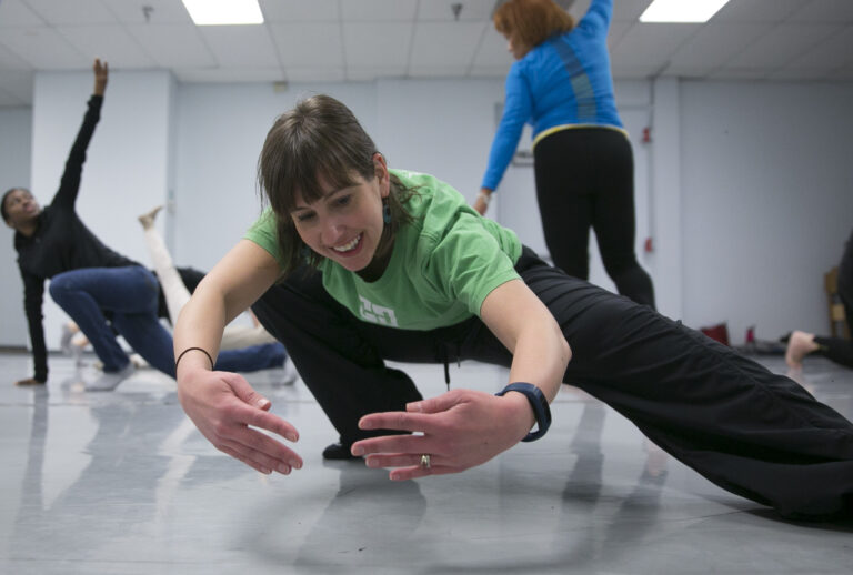 Dancer leaning towards the right in a squat and holding arms in a circular position. She's wearing a green tshirt and black pants.