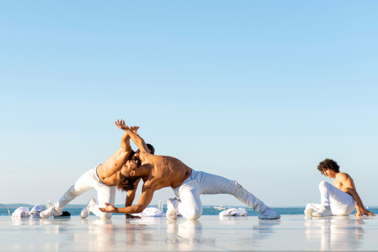 On a wet sandy beach in front of a clear light blue sky, three male dancers in white pants dance together. The two closest to the camera mirror each other back to back, arching backward as they lunge back and rest on their right knees for support, chests turned up toward the sky.
