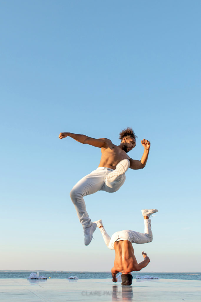 On a wet sandy beach in front of a clear light blue sky, three male dancers in white pants dance together. In the foreground, Houssni Mijem is mid-leap with arms pumped and legs extended. In the background, another dancer spins on his head.