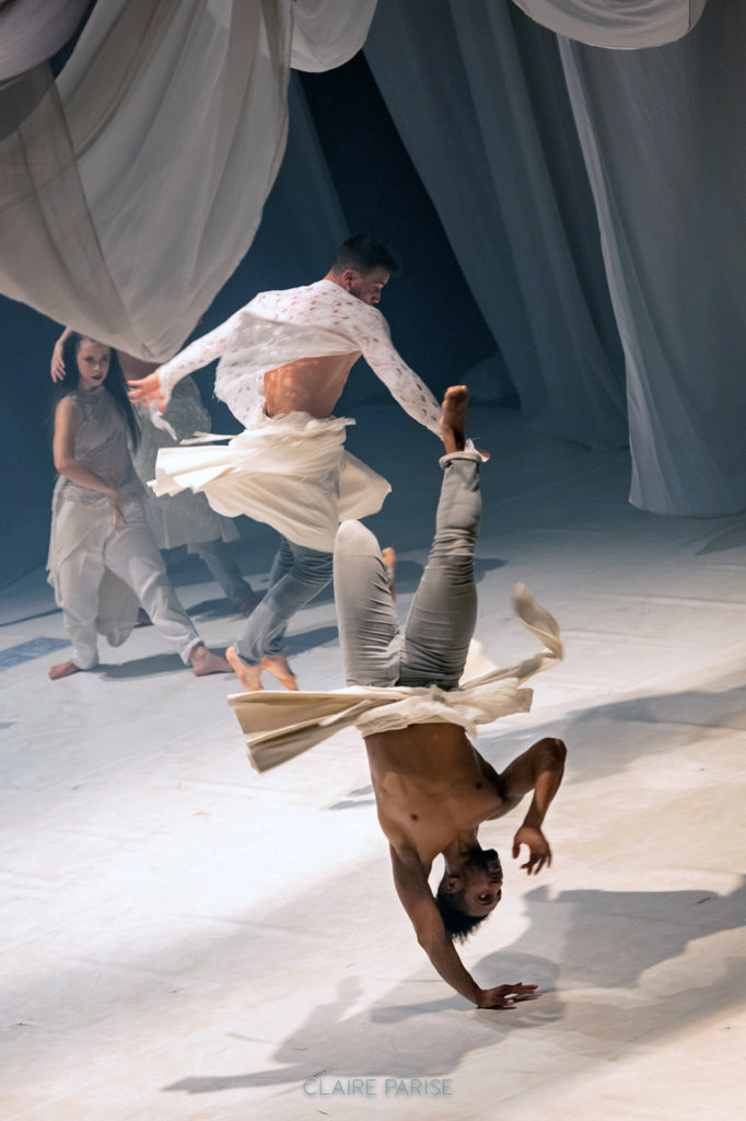 On a stage with contrasting lighting and gray decorative curtains, male and female dancers in flowy white and gray clothing dance together. In the foreground, Houssni Mijem is shown mid-head spin, while another male dancer jumps behind him.