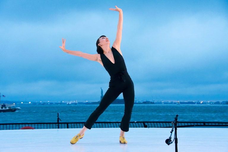 On an outdoor stage in front of a blue sky and large body of water, Demi Remick poses energetically, arms and head lifted up in a slight lunge. She wears a black jumpsuit and tan heeled tap shoes.