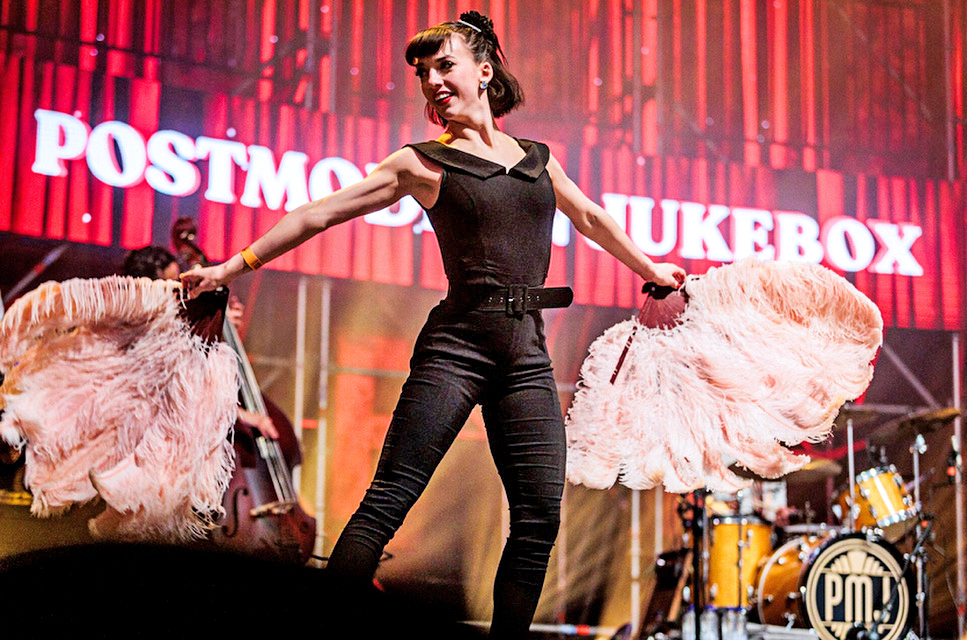 In front of a large red curtain and a sign reading "Postmodern Jukebox," Demi Remick performs a tap dance holding light pink feather fans. She wears a black belted jumpsuit.