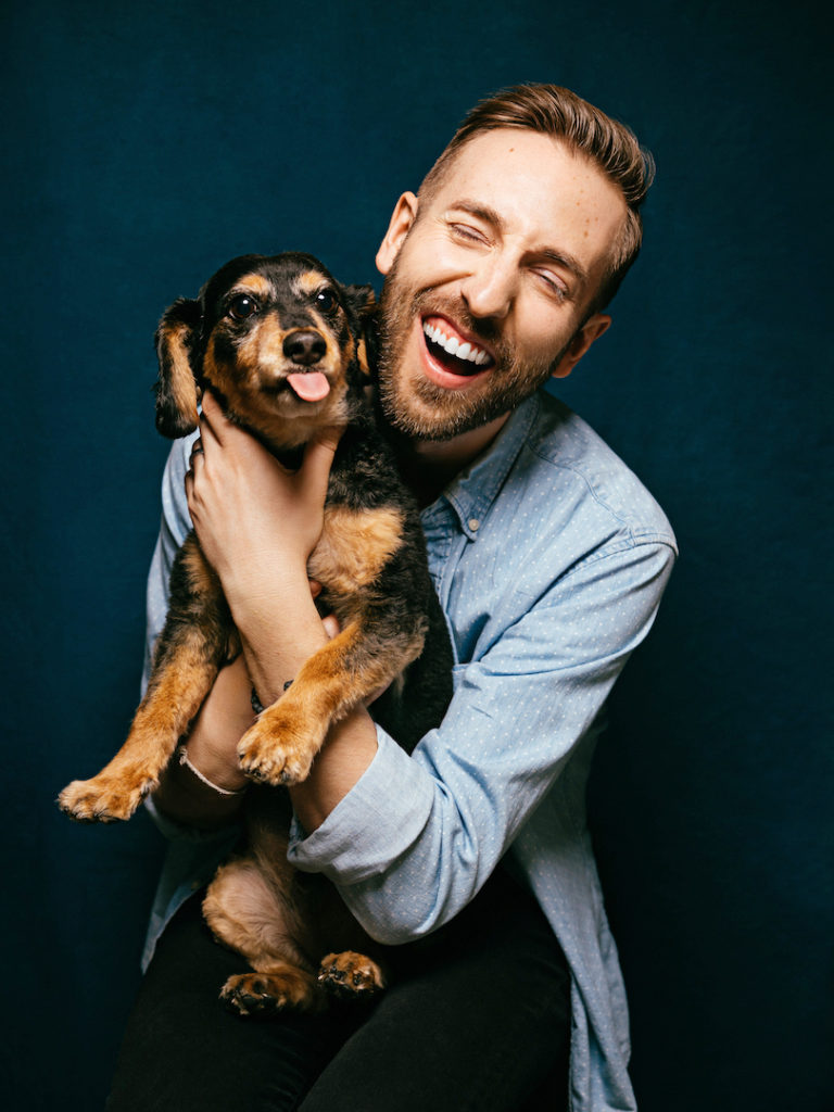 A young man in a light blue button down shirt smiles with his eyes closed, hugging a brown and black puppy.