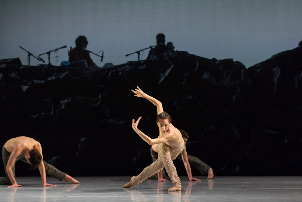 Onstage in front of an orchestra's silhouette and a bluish gray backdrop, Ilaria Guerra wears a nude unitard and twists her torso in a deep lunged tendu croise derriere, her arms swiping up to the right. Other dancers do floorwork around her.