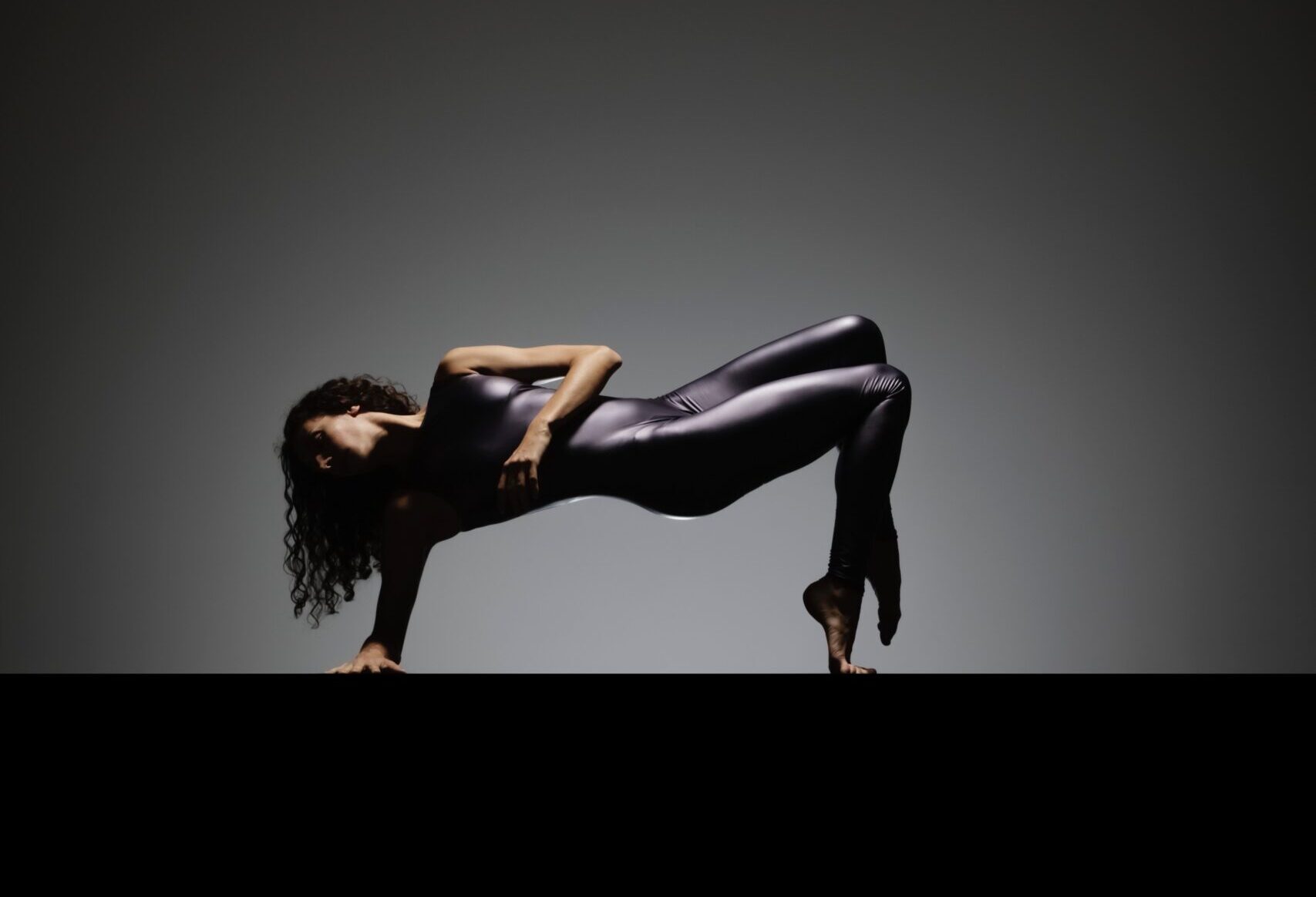 In front of a gray backdrop, Ilaria Guerra does a stylized bridge pose in a shiny grayish-purple unitard. Her long dark curly hair is down, and as she uses her right arm and toes on forced arch for support, she twists toward the camera with her left arm crossing over her chest.