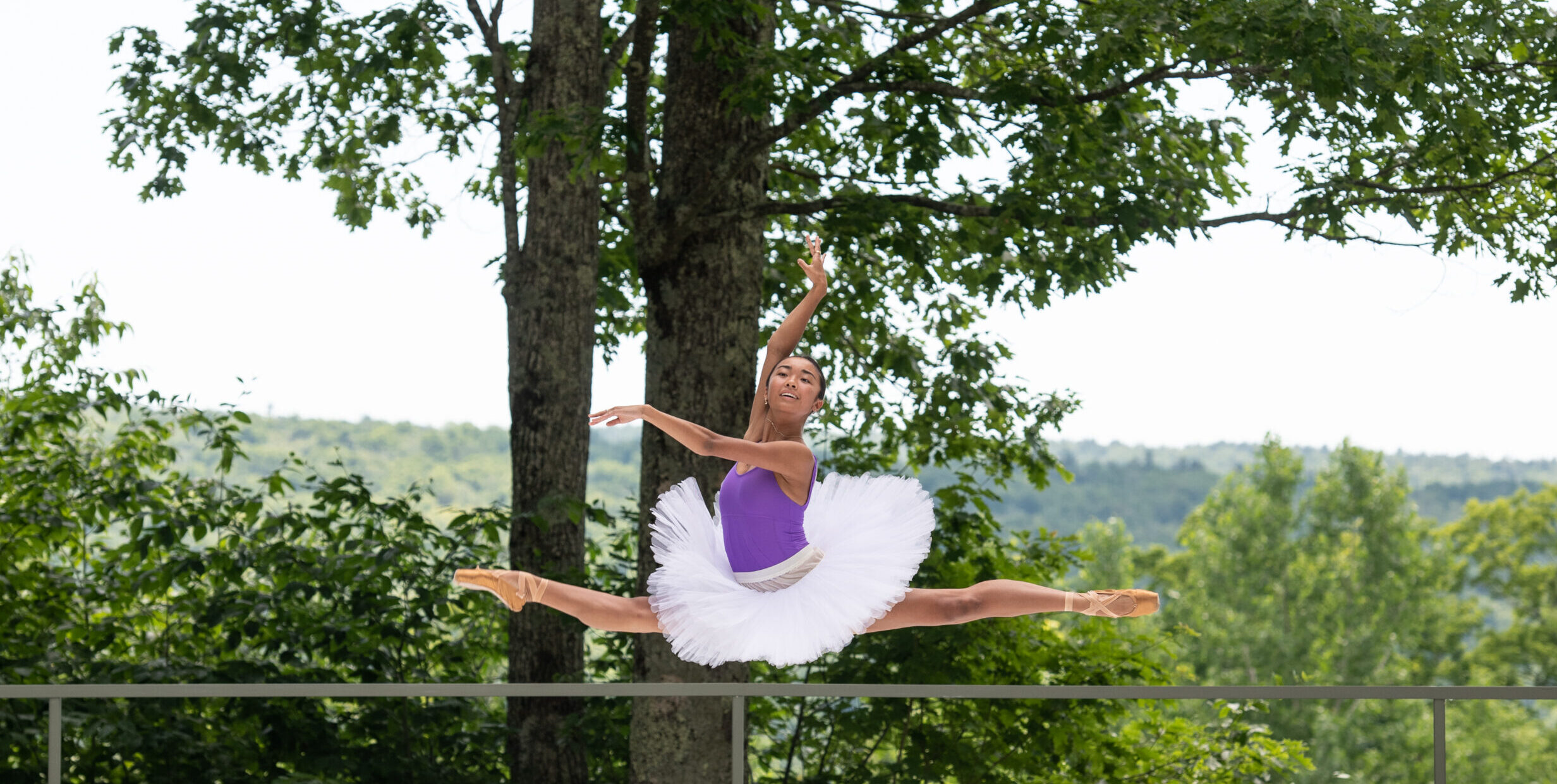 At an outdoor performance space, a young female dancer in a purple leotard, white pancake tutu and pointe shoes does a flying saut de chat in front of trees.