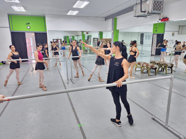 Marena Pérez wears a black tank, leggings, and sneakers standing at a barre at the front of a ballet studio teaching a group of teenage girls.