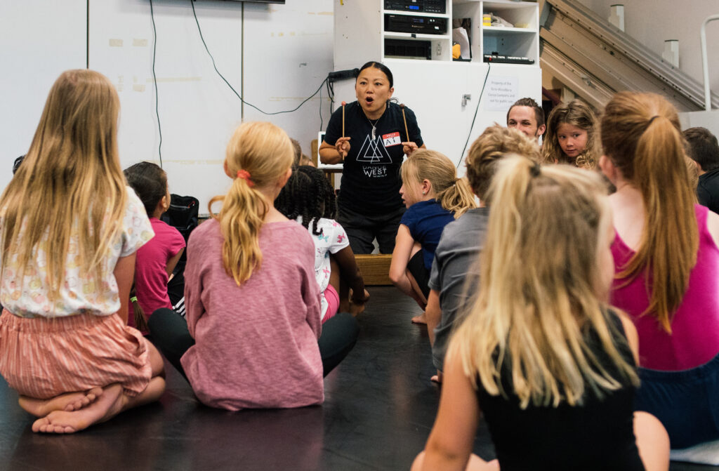 In a dance studio, a female instructor kneels facing a group of young dance students, who kneel facing her and watch as she animatedly plays a percussive instrument.