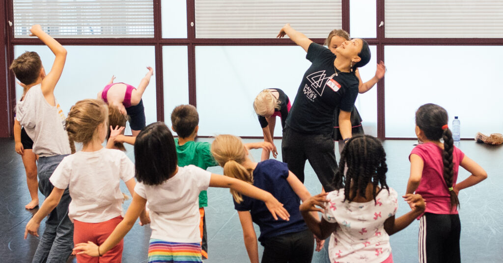 In a bright dance studio, a female dance teacher leads a large class of young dance students. They all wear colorful clothing and move together making sweeping movements with their arms.