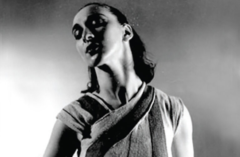 In this black and white photo, Anna Sokolow is shown chest-up in a layered fabric top with short sleeves. Her short hair is half-pulled back, and she leans back slightly as she tilts her chin up and looks over her right shoulder.