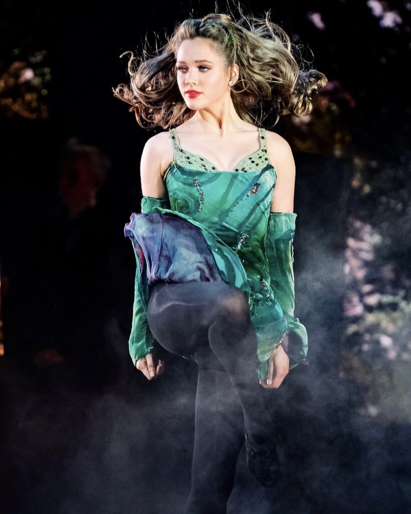 In front of a black backgound lit with textured lights, a female Irish dancer is shown mid-jump as her kelley green costume dress flies around her, her hair lifted around her face.
