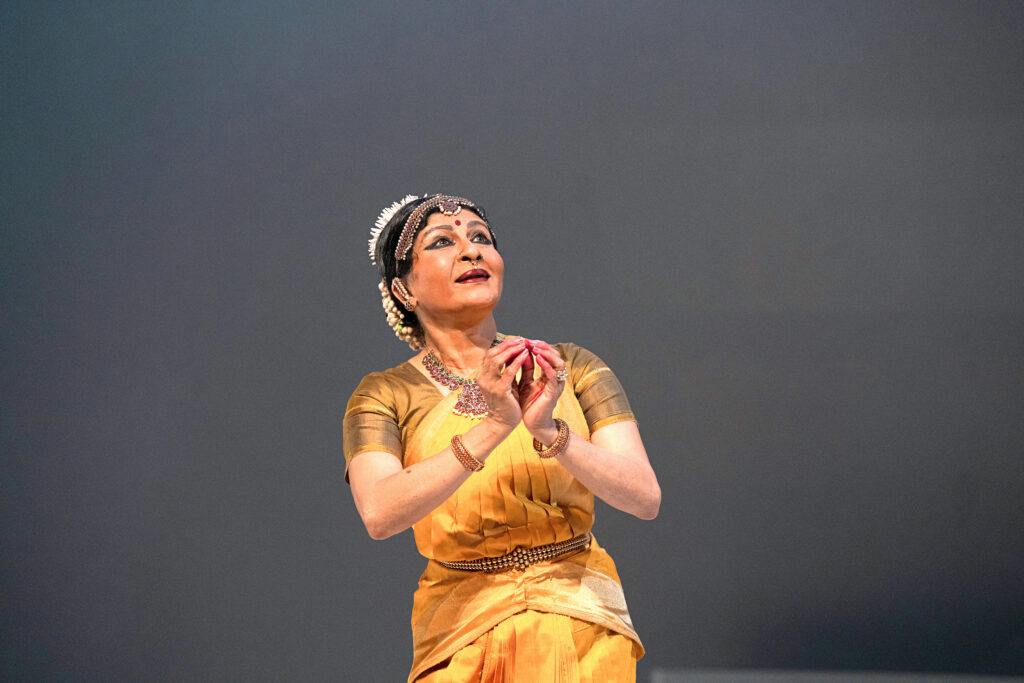 Dancer Ranee Ramaswamy performs bharatanatyam on stage wearing a yellow costume, with her eyes looking upward.