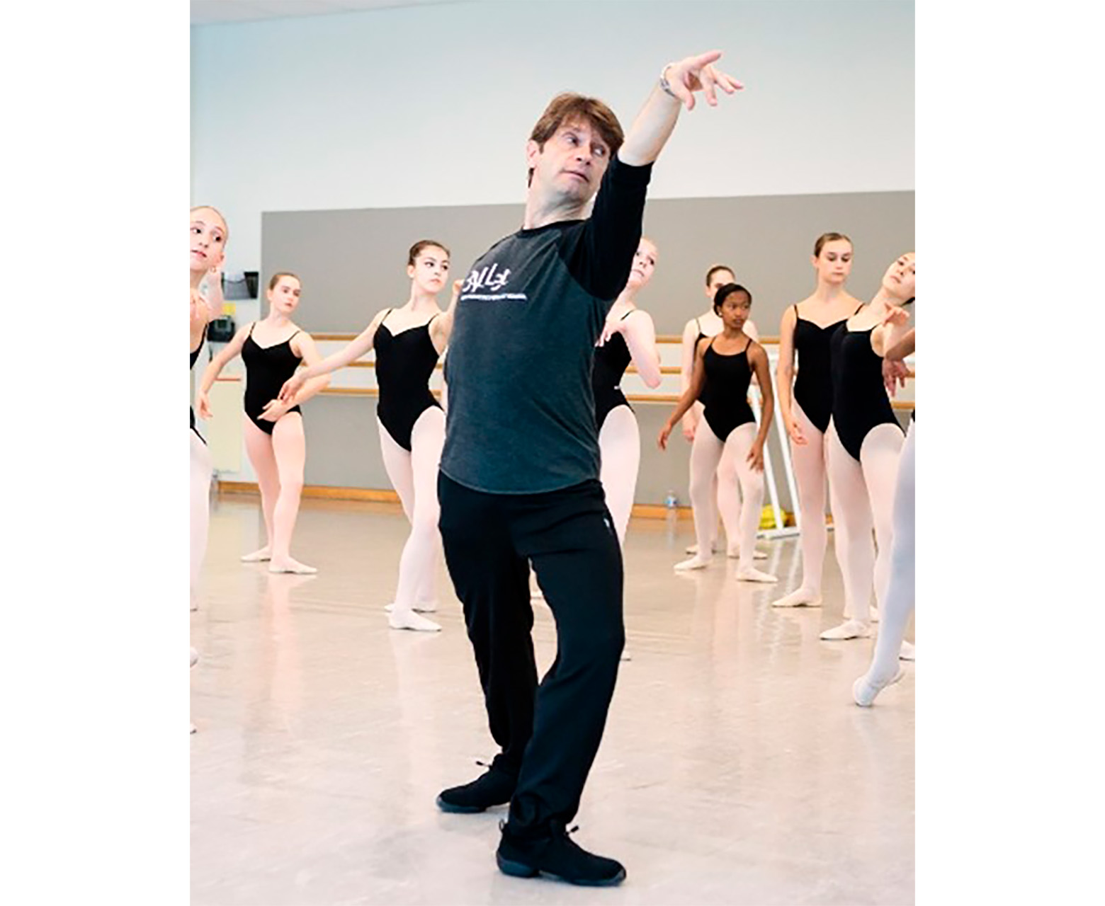 Patrick Armand demonstrates for a ballet class. He is in a fourth position lunge, downstage arm reaching long over his front leg, head inclined toward the front of the room as he illustrates épaulement. He wears a San Francisco Ballet branded long sleeve shirt. Behind him, young dancers in black leotards and pink tights variously watch and copy him.