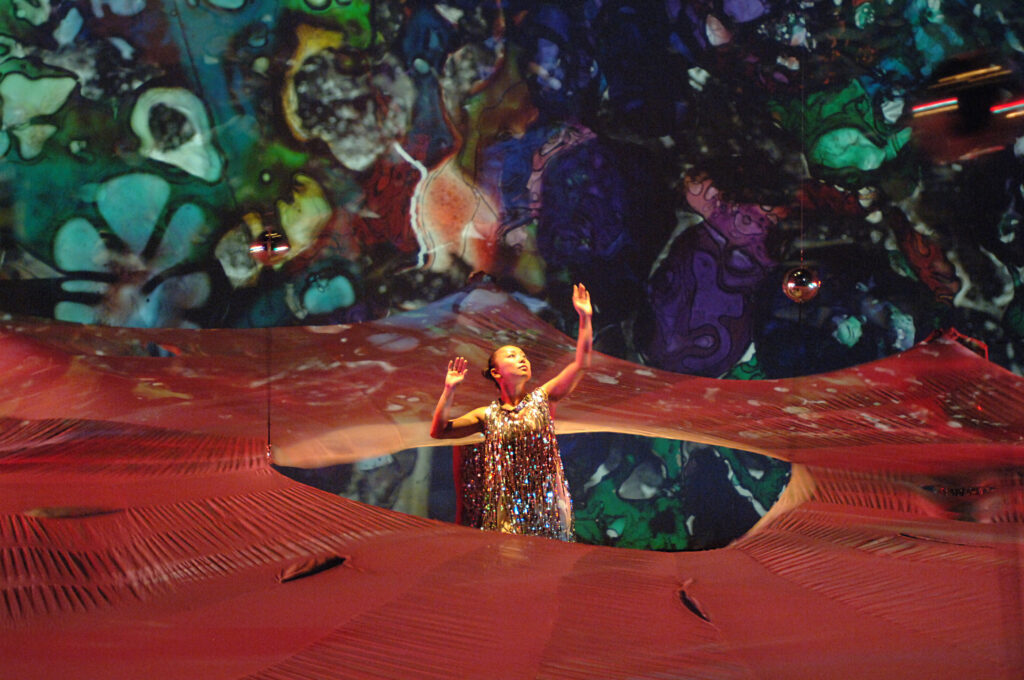 In an interactive performance space covered with projections of glass artwork, a female dancer in a shimmering dress reaches upward in wonder as she stands in a waist-high structure mimicking a glass-blown flower.