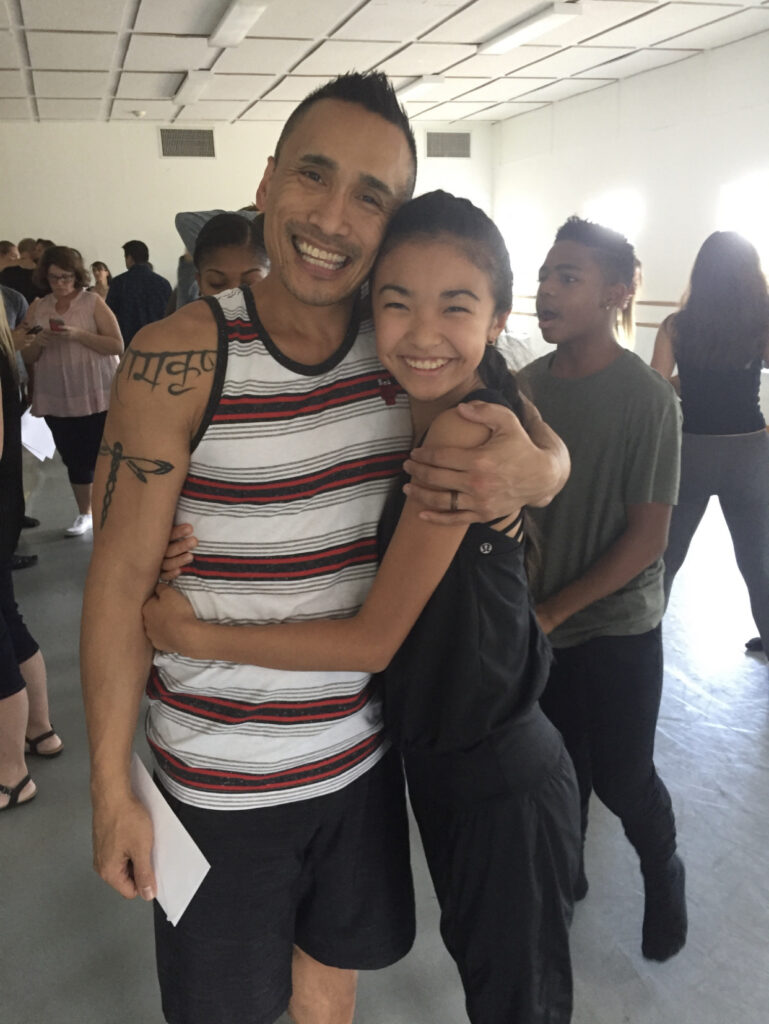 In a dance studio, a male teacher and a female student hug and smile at the camera.