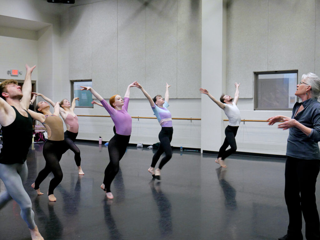 A group of college students in colored leotards and black tights dances across a studio floor with their arms in a Paul Taylor "v" behind them.