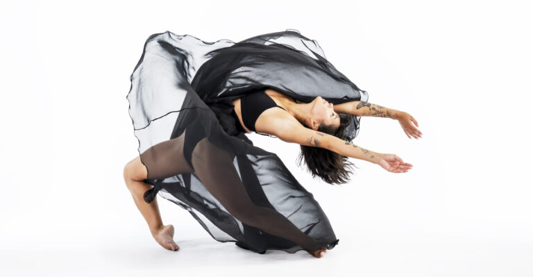In front of a bright white background, a female dancer with floral forearm tattoos does a dramatic arch backward on forced arch, her knees bent in a wide double-attitude position. She reaches her arms overhead, parallel to the floor. as the black tulle of her skirt flies around her.