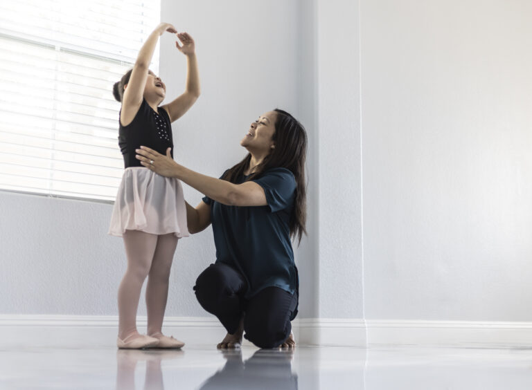 Dance teacher kneels on the floor, adjusting a young girl's fifth position arms.