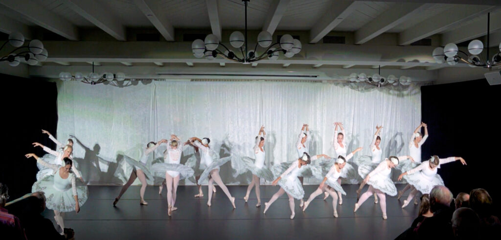 A corps de ballet of adult ballet students perform a scene from "Swan Lake" in a studio theater. They stand in plié on their right legs with their left leg behind them in tendu derriere and create various formations. They all wear white tutus with white long-sleeved leotards, pink tights and ballet slippers, and feathered headpieces.