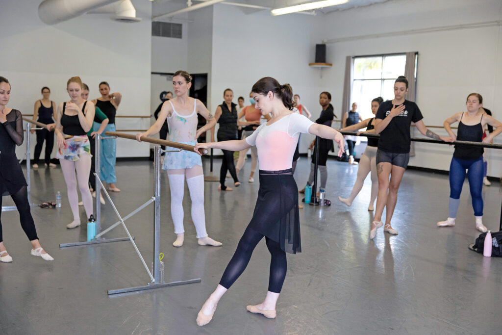 Kathryn Morgan, wearing a pink leotard, black ballet skirt, black tights and ballet slippers, demonstrates a tendu devant while holding onto the end of a portable barre in a crowded ballet studio. She is surrounded by adult ballet students in various dancewear, who stand at their places at the barres and watch her intently.
