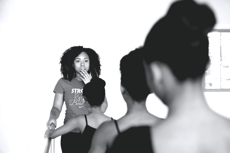 Aa black and white image of Karisma Jay standing at the barre in a t-shirt with the backs of three young dancers facing the viewer.