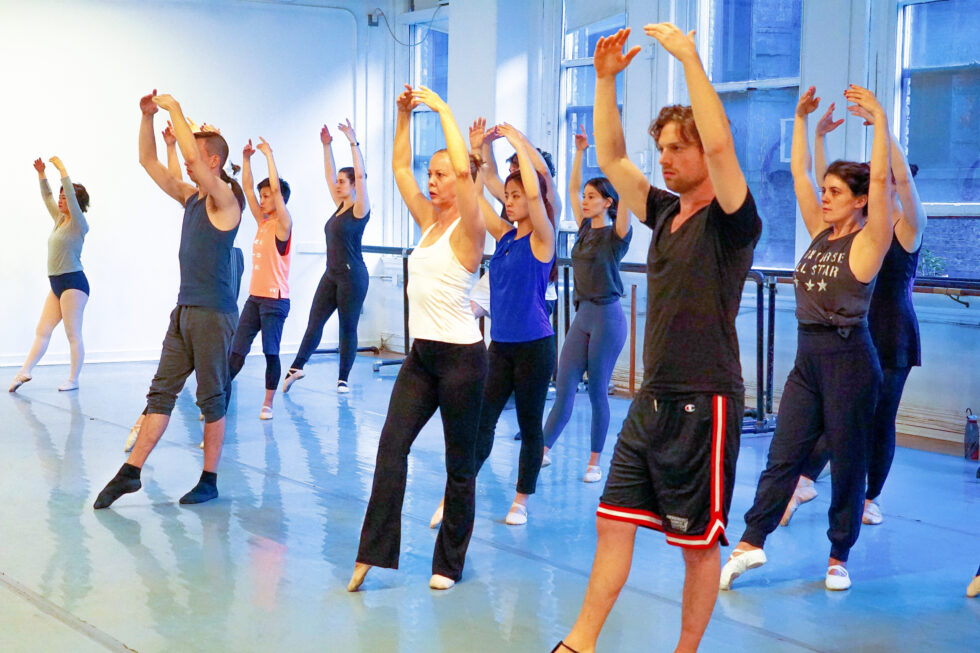 Kat WIldish stands among her adult students in her ballet class and demonstrates a tendu devant en face with her arms in high fifth. Her male and female students, looking focused, standing in three lines and follow along with her. Wildish wears a white tank top, black yoga pants and ballet slippers. Her students wear various workout clothing (leggings, shorts, and T-shirts) and ballet slippers, as well.