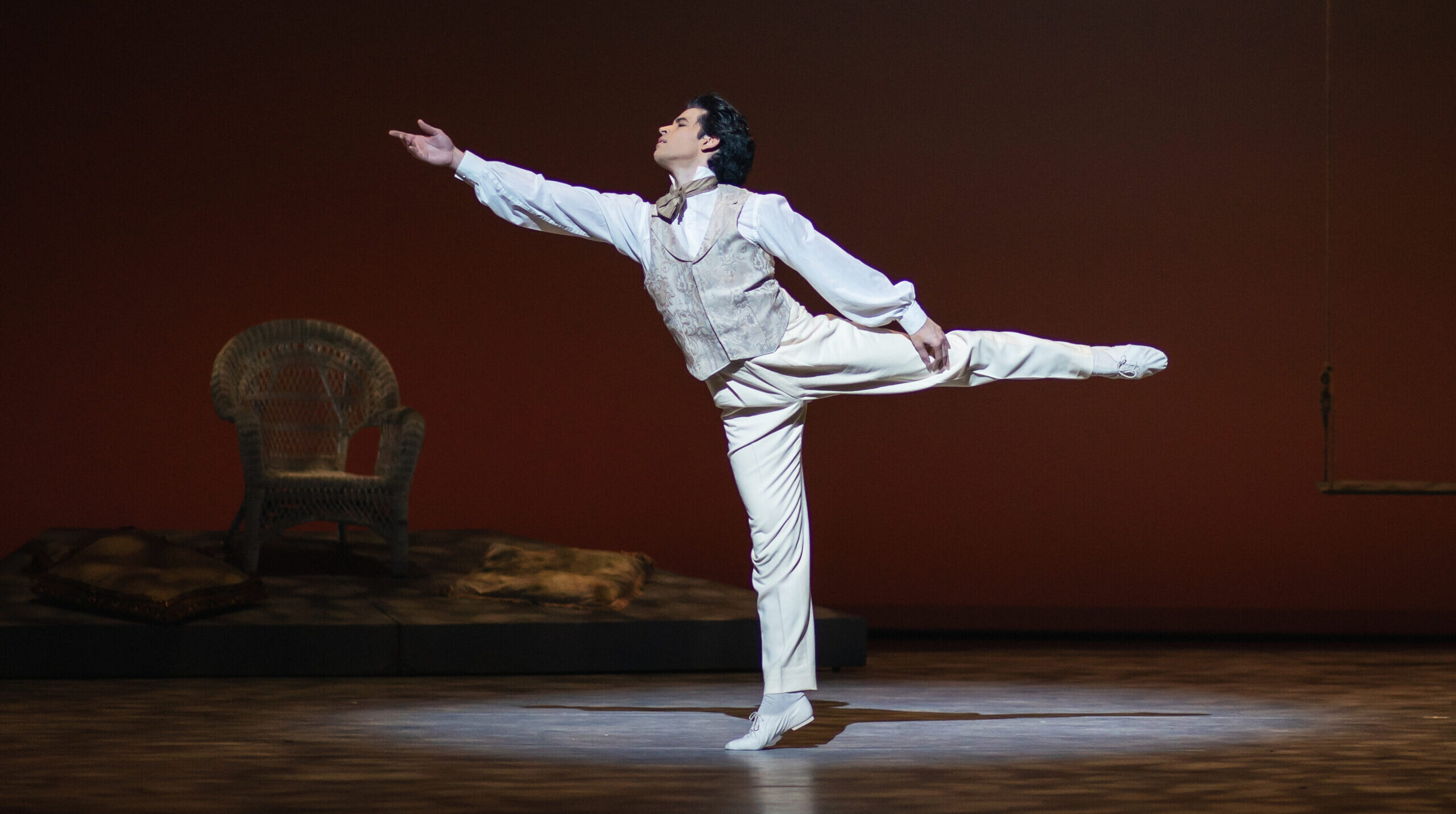 Onstage, a male dancer in a white, cream, and light tan suit costume does an arabesque onstage, his right arm reaching forward with longing.