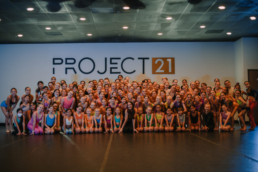 In a dark dance studio with mood lighting, a large group of dance students pose for a group photo with their dance teacher in front of a white wall with "Project 21" painted in large block letters.