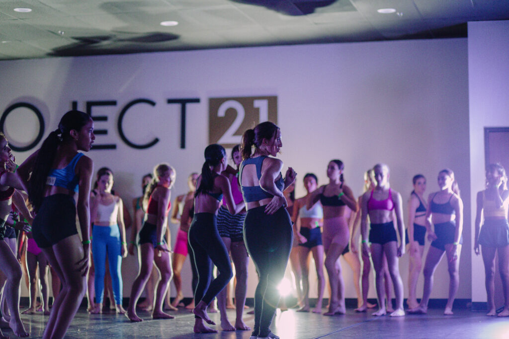 In a dark dance studio with colorful mood lighting, a female dance teacher in athletic clothing demonstrates a dance combination for a group of young students, who follow along with her movements behind her.