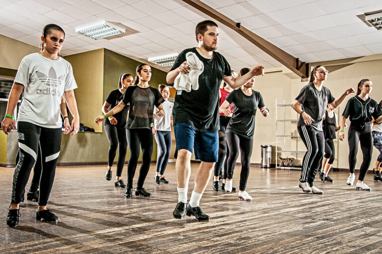 Anthony Morigerato wears a t shirt and shorts and stands at the front of a class of teenage tap dancers, demonstrating as they follow along.
