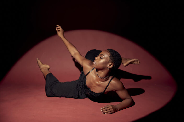 In front of a dark red background, a Jada Clark poses on the floor, lit by a spotlight. She wears a black jumpsuit and lies on her side, her legs each in a stylized attitude position, as she twists up to the right and reaches her right arm up on the diagonal, using her left elbow for support on the floor.