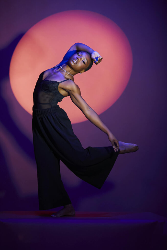 In front of a deep purple backdrop, Jada Clark is lit in a spotlight that turns the wall behind her pinkish-red. She dances in a long black jumpsuit, her left leg lifted in a parallel attitude behind her, her left arm reaching back to touch her foot, and her right arm folded over top of her head as she bends back slightly.