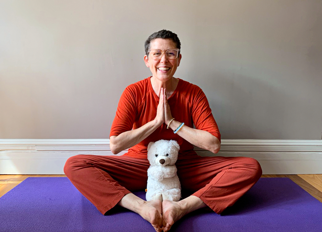 Hetty King wears all red and sits on a purple yoga mat with her hands in a prayer pose, a white teddy bear positioned between her legs. She's smiling widely.