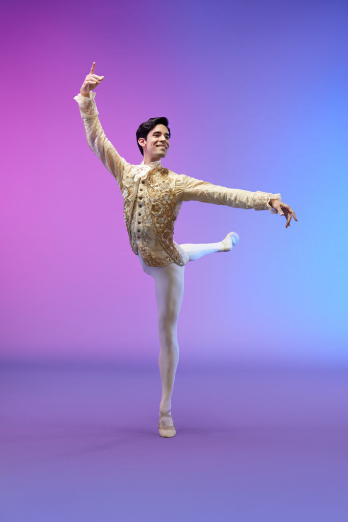 In front of a multicolored pink, blue, and purple background, a male dancer in a gold and white prince costume does an attitude derriere balance, his arms held high in third position, and his head looking over his left arm. He smiles generously.
