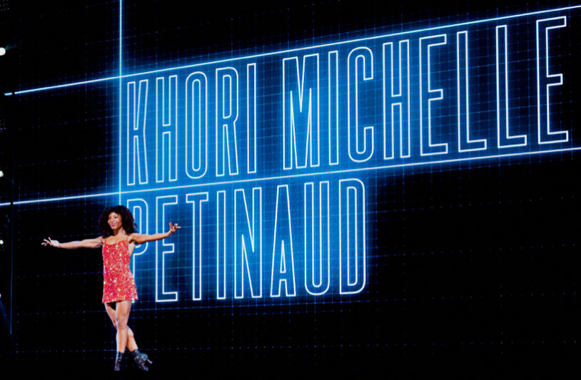 A female dancer in a short sparkly red dress and Laduca dance books poses confidently in front of a large blue neon sign with her name "Khori Michelle Petinaud," in large block letters.