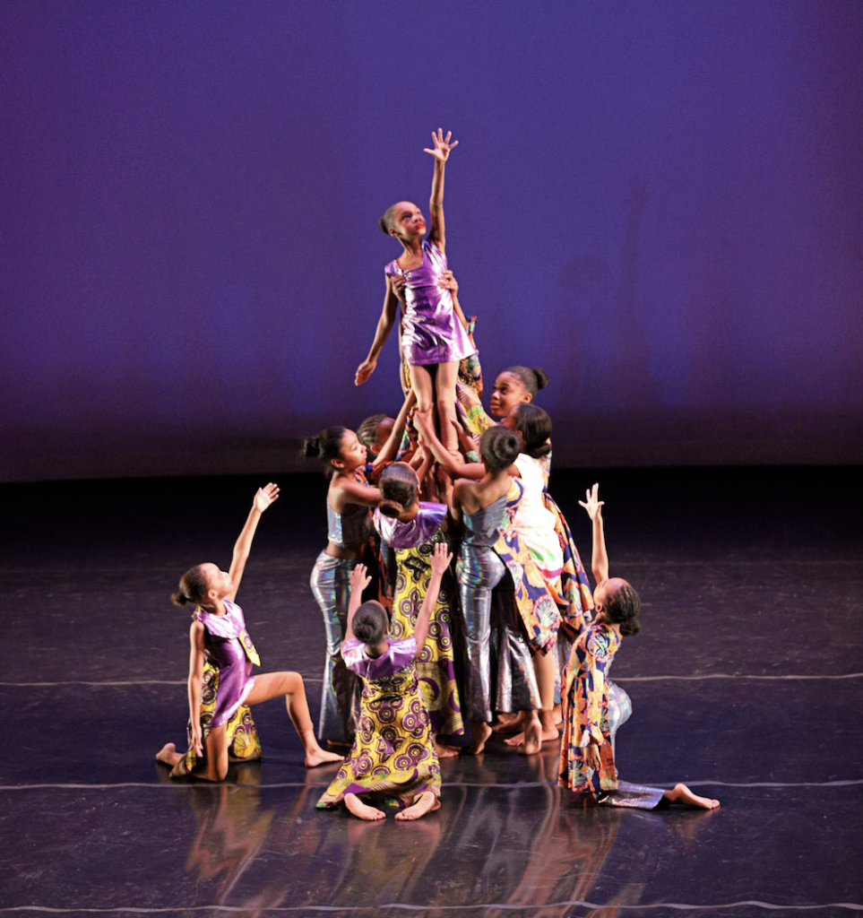 A group of young students in purple, gold, and gray costumes performs onstage in front of a dark purple background forming a huddle and lifting one dancer into the air.
