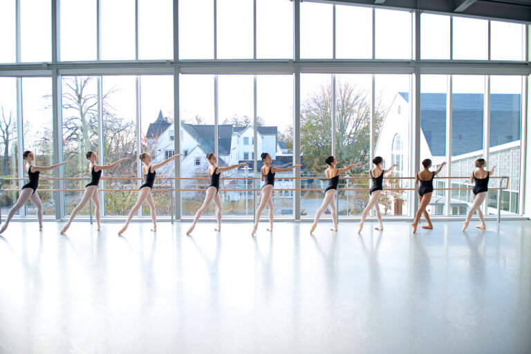 A group of young ballet dancers stand at the barre in black leotards and pink tights with one leg extended behind them in front of large glass windows.