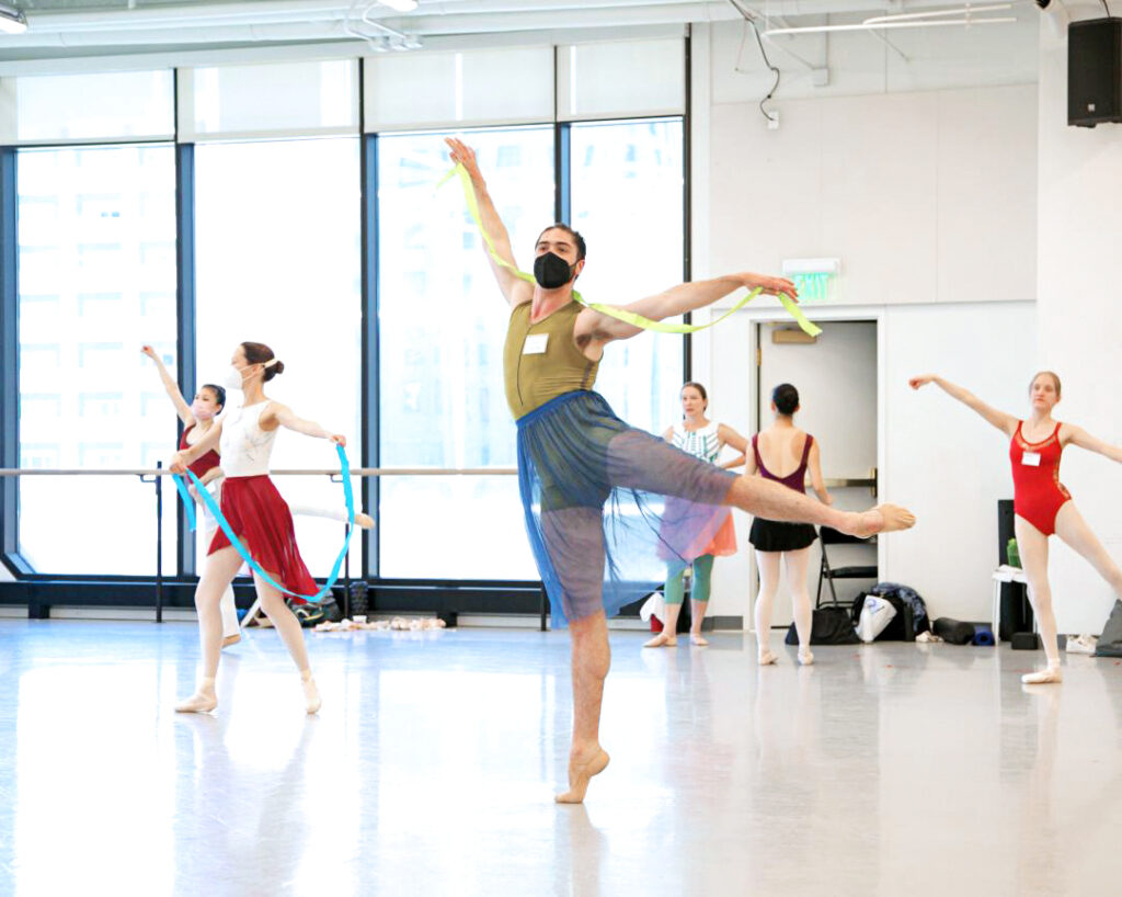 A tall adult ballet student practices a first arabesque and looks out towards the front of the studio. The dancer wears a green biketard, a sheer, knee-length blue skirt, tan ballet slippers and a black face mask, and holds a long green ribbon in both hands, which around their shoulders.