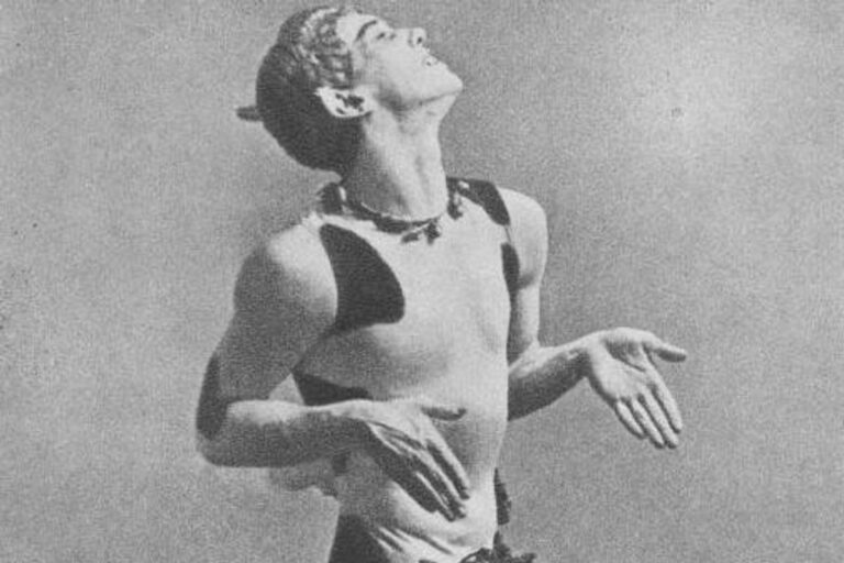 A black and white photo shows a close-up of Vaslav Nijinsky in costume for "Afternoon of a Faun." He wears a satyr costume and makes a stylized pose with bent elbows and wrists, completely profile, and lifts his chin upwards with a smiling grimace.