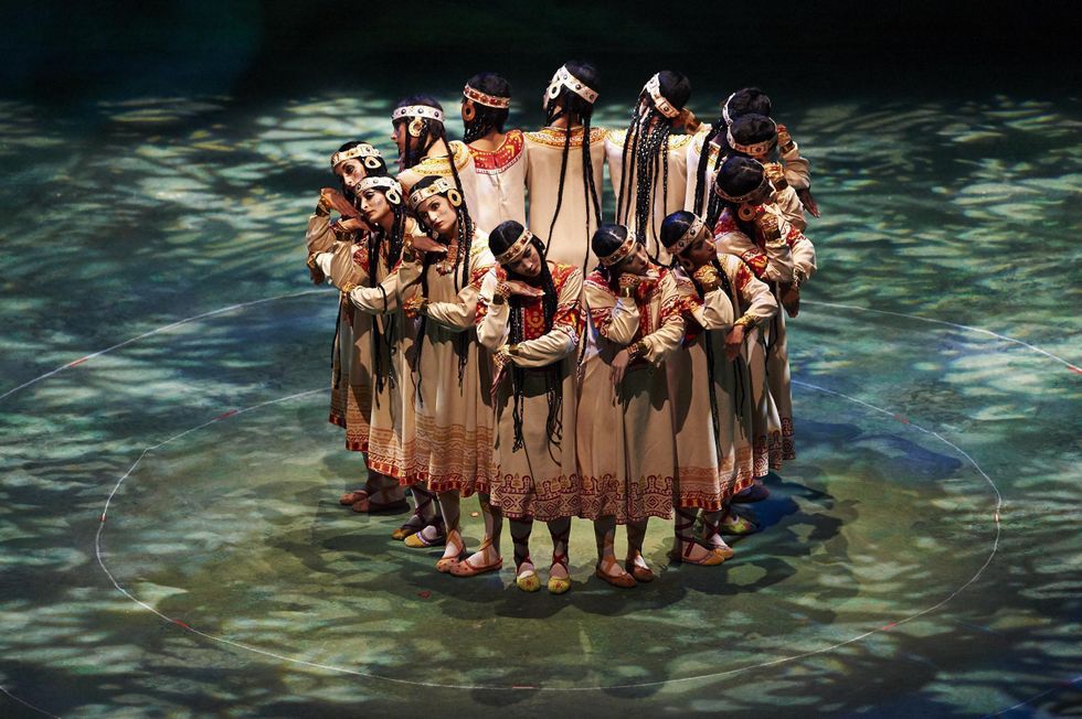 Members of The Joffrey Ballet perform a reconstruction of Nijinsky's "Rite of Spring." They wear outfits reminiscent of indigenous communities, with long braids, and form a tight circle linking arms. The stage is lit with a textured greenish light.