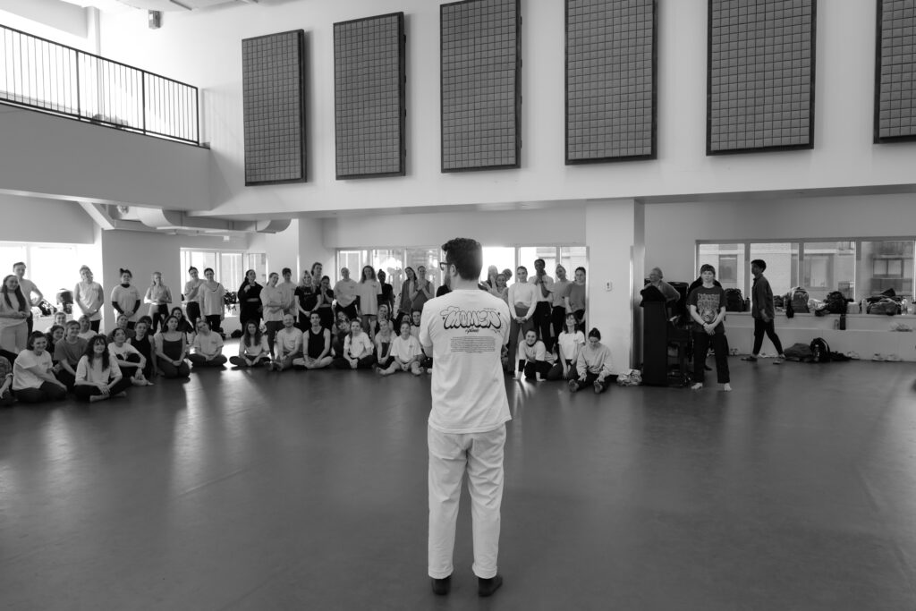 Black and white photo of Al Blackstone from the back speaking to a large group of dancers.