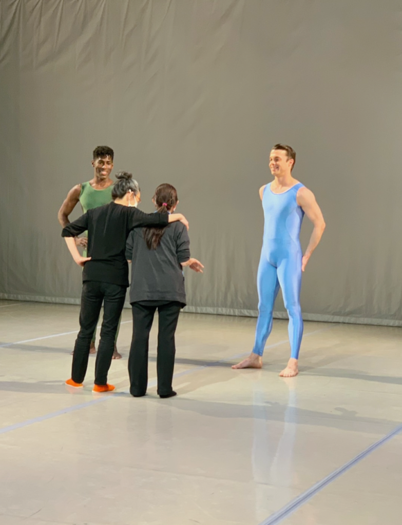 Miki and Susan have their arms around each other. We see them from the back, as they face two dancers in unitards on a stage. 