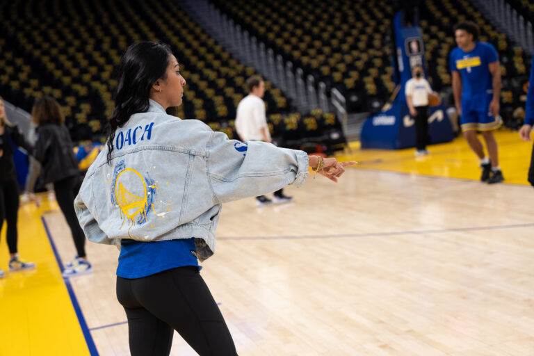 Sabrina Ellison faces her side toward the camera wearing a custom jean jacket that says "COACH" with the Golden State Warriors logo. She points toward the court as she speaks to her dance team.