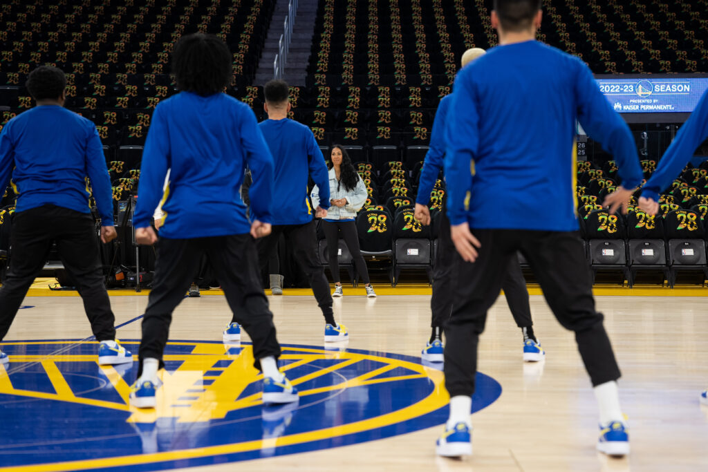 On the Golden State Warriors' court, the dance team's male dancers rehearse a routine with their coach, Sabrina Ellison, watching them.