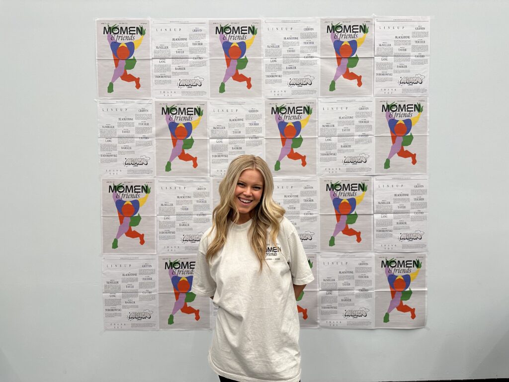 Haley Hilton poses in a white t shirt in front of a wall covered in Momen posters.