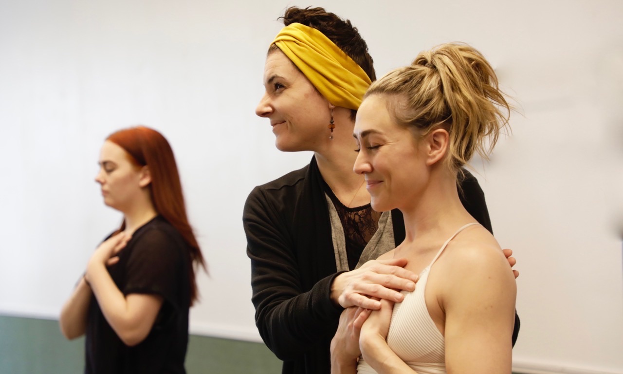 Chryssie Whitehead, wearing a dark sweater and wide yellow headband, stands in a studio with her hand on aa student's upper chest, helping her breath.