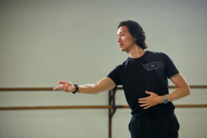 Runqiao Du gestures with his arms in front of a ballet barre, wearing a dark t-shirt.