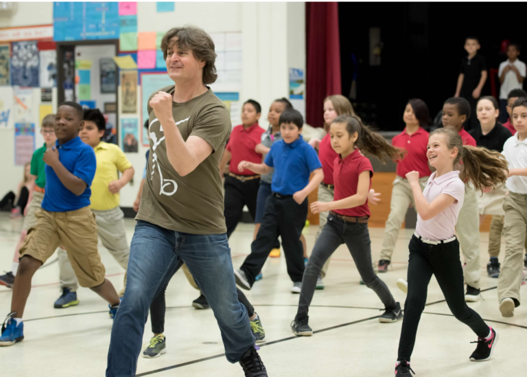 A teacher in a t-shirt and jeans leads a group of elementary school aged dancers in a school auditorium.