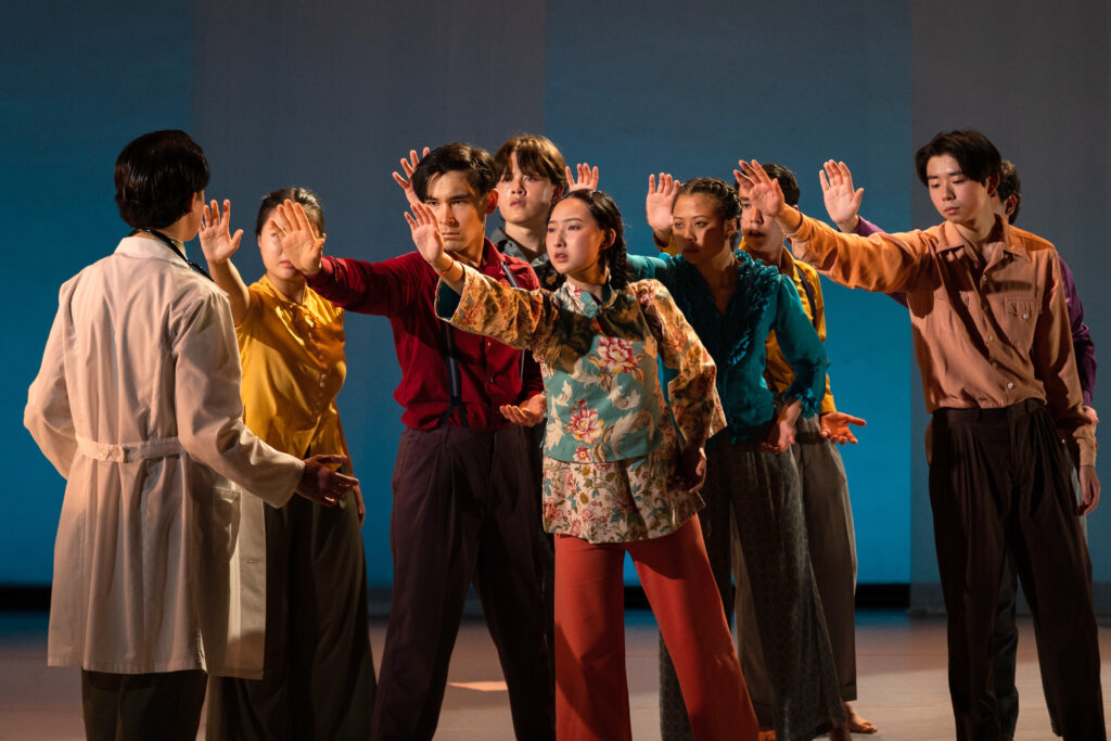 Performance photo of a group of eight Asian dancers standing with one hand out in protest towards a figure in a doctor's coat.